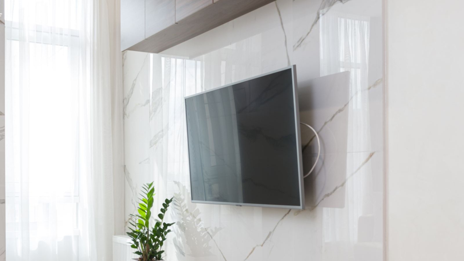 Telewizor zawieszony na jasnej ścianie wykończonej płytą imitującą marmur, obok zasłony i roślina